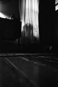 a black and white photo. a figure shrouded by a curtain sits hunched in the dark.