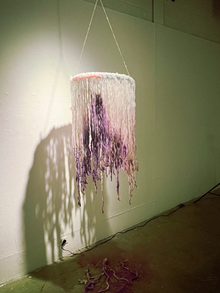 a photo of a sculpture. Strands hang down from a circular rim like tentacles, the ends dyed purple.